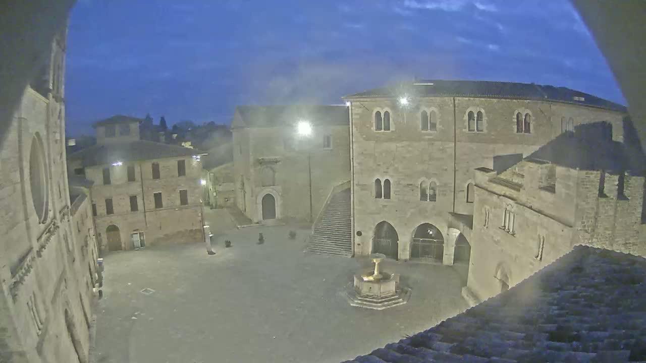 A nighttime view of a town square, with stone buildings surrounding a central fountain under a twilight sky.