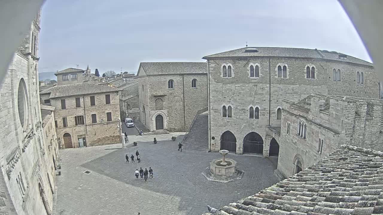 A town square with several stone buildings, a fountain, and a few people, under a clear sky.