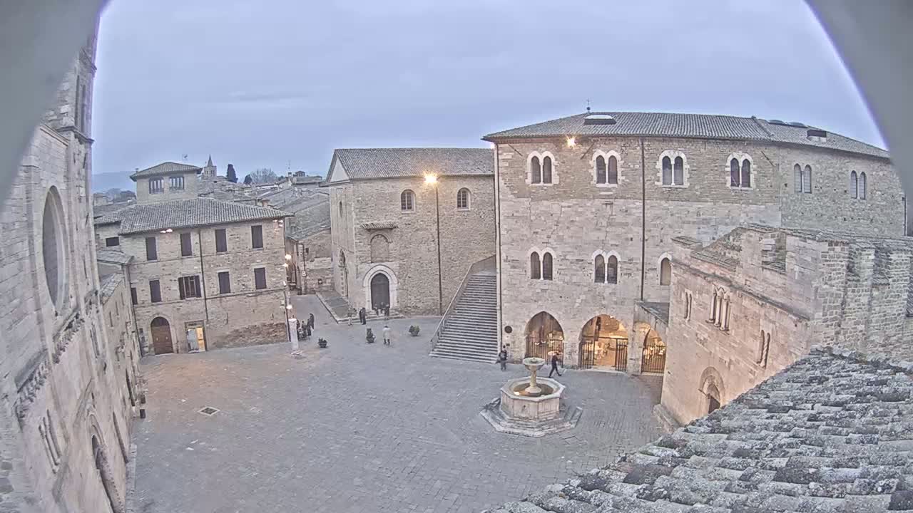 A stone plaza with a fountain is surrounded by medieval-style buildings under a cloudy, twilight sky.