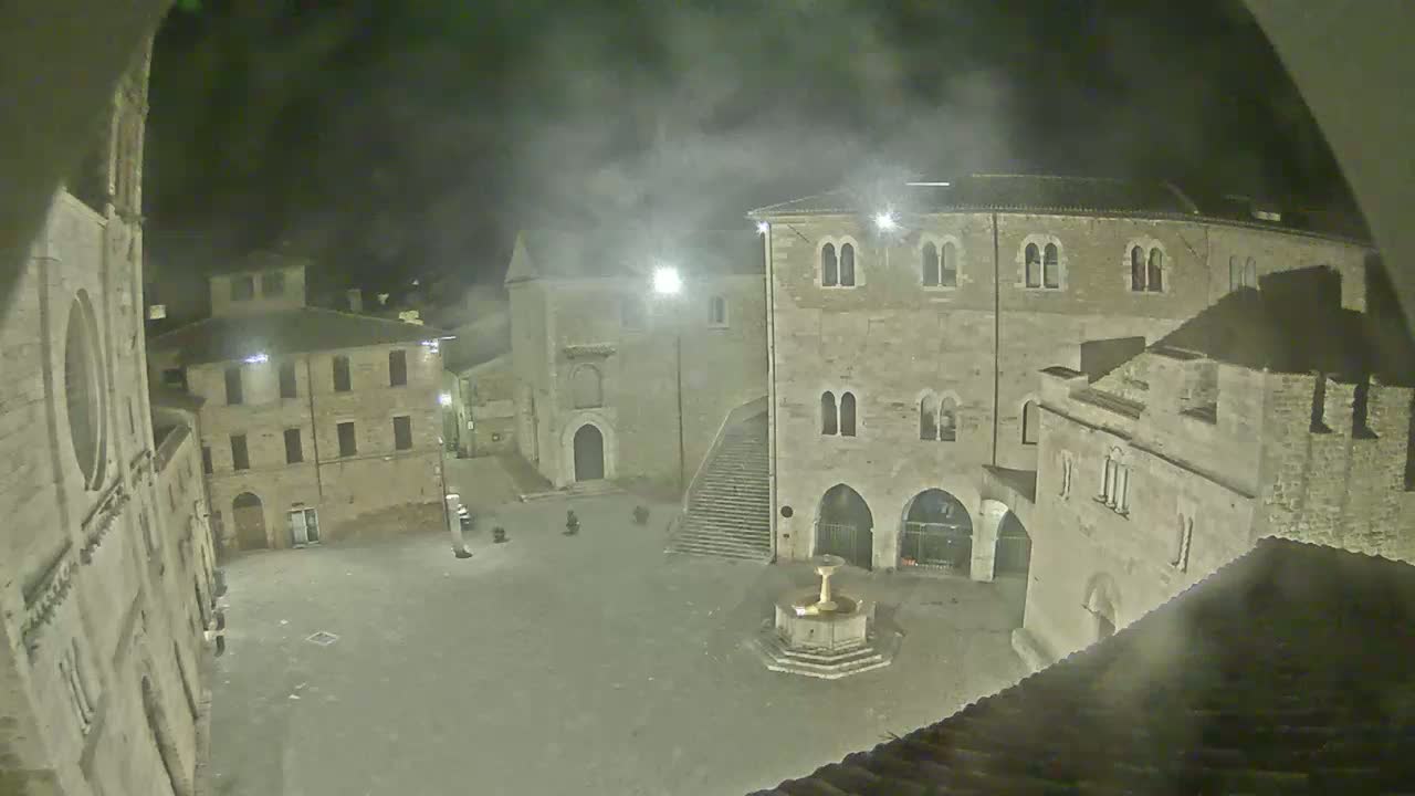 A nighttime view of a stone town square, illuminated by streetlights, featuring a fountain and several historic buildings under a hazy or foggy sky.