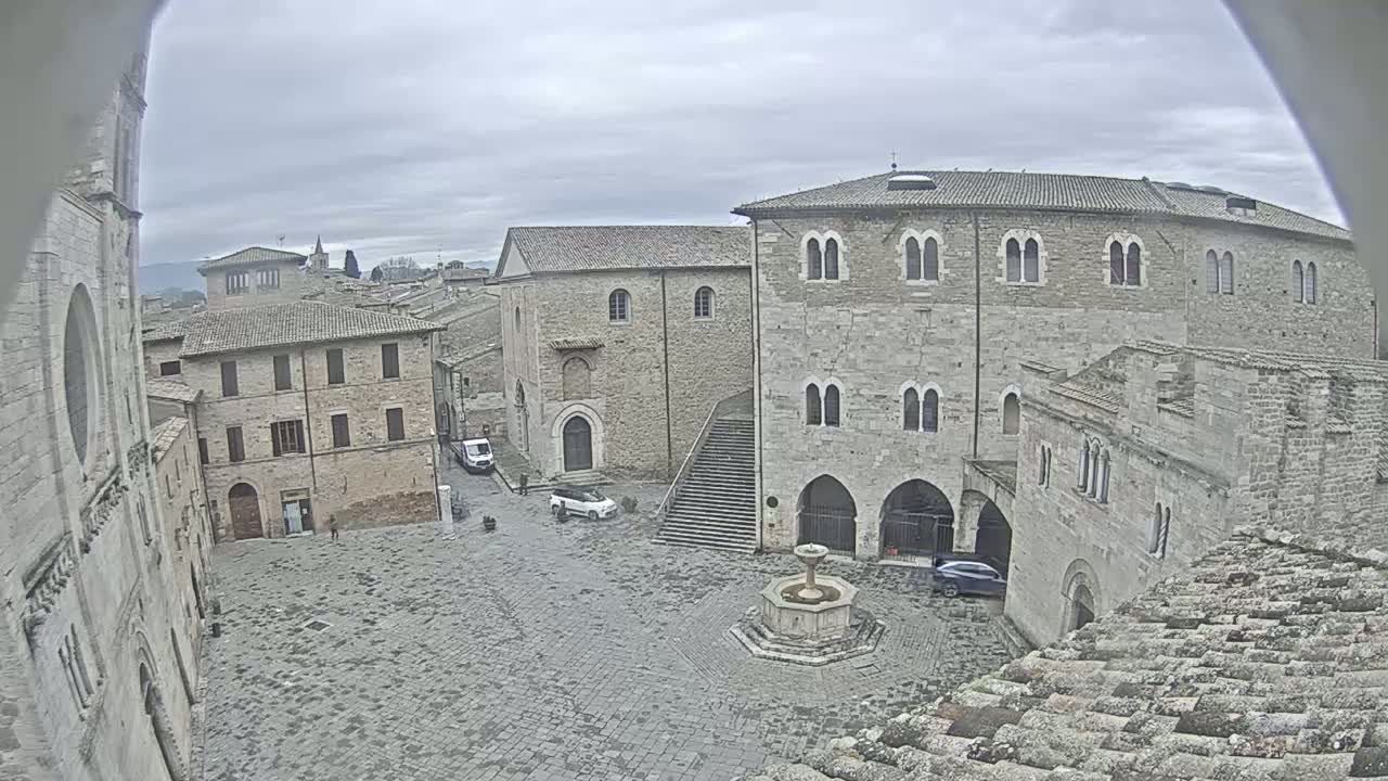 A stone plaza with a fountain is surrounded by medieval-style buildings under an overcast sky.