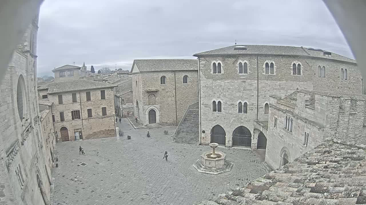 A stone plaza with a fountain in the center, surrounded by old stone buildings, under an overcast sky.