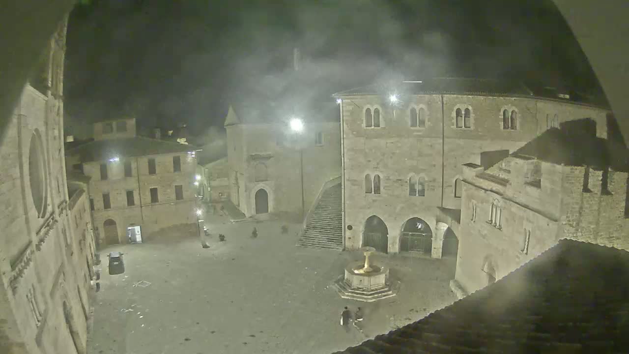 A nighttime, hazy view of a town square with old stone buildings surrounding a fountain, several people visible in the square.
