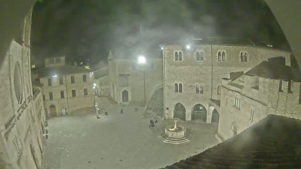 A nighttime, hazy view of a town square surrounded by ancient stone buildings, featuring a fountain in the center.