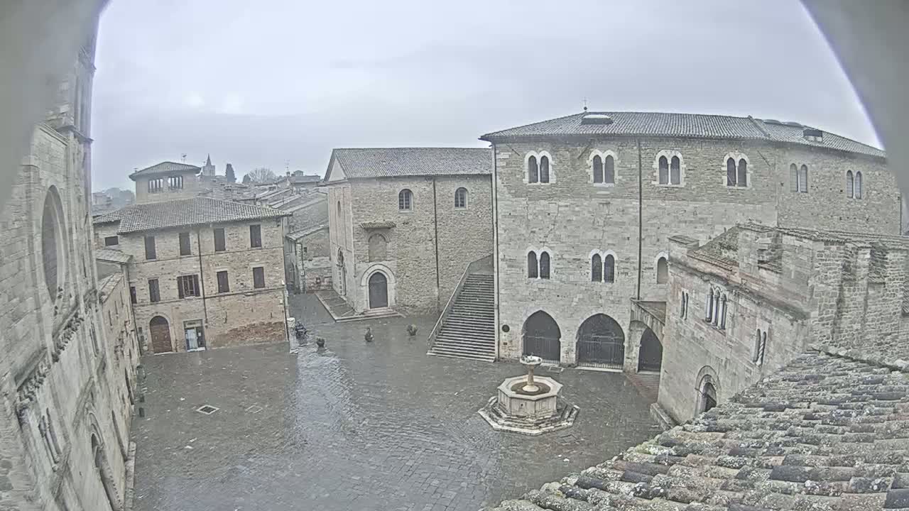 A wet, stone plaza in a historic town is viewed from above, featuring several aged buildings, including a church, surrounding a central fountain.
