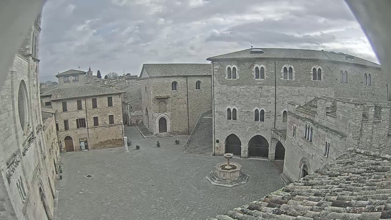 A stone plaza with a fountain is surrounded by medieval-style buildings under a cloudy sky.