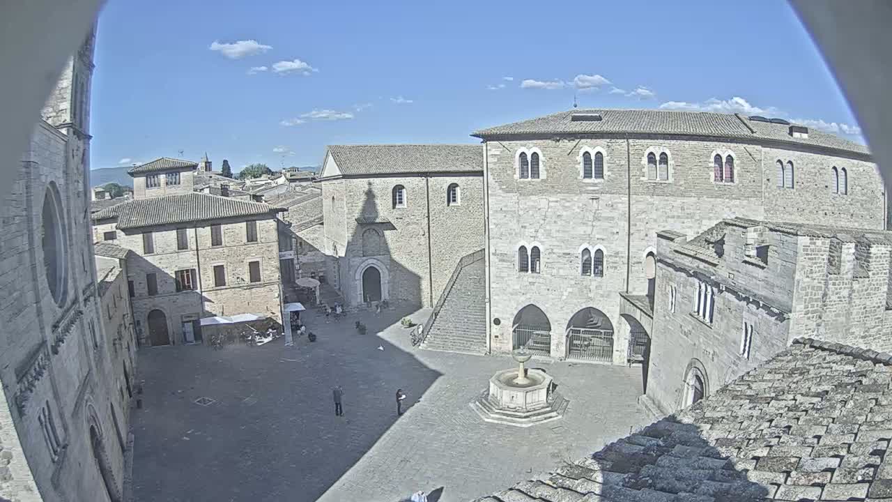 A sunny day reveals a stone plaza in a medieval town, centered by a fountain and surrounded by historic buildings.