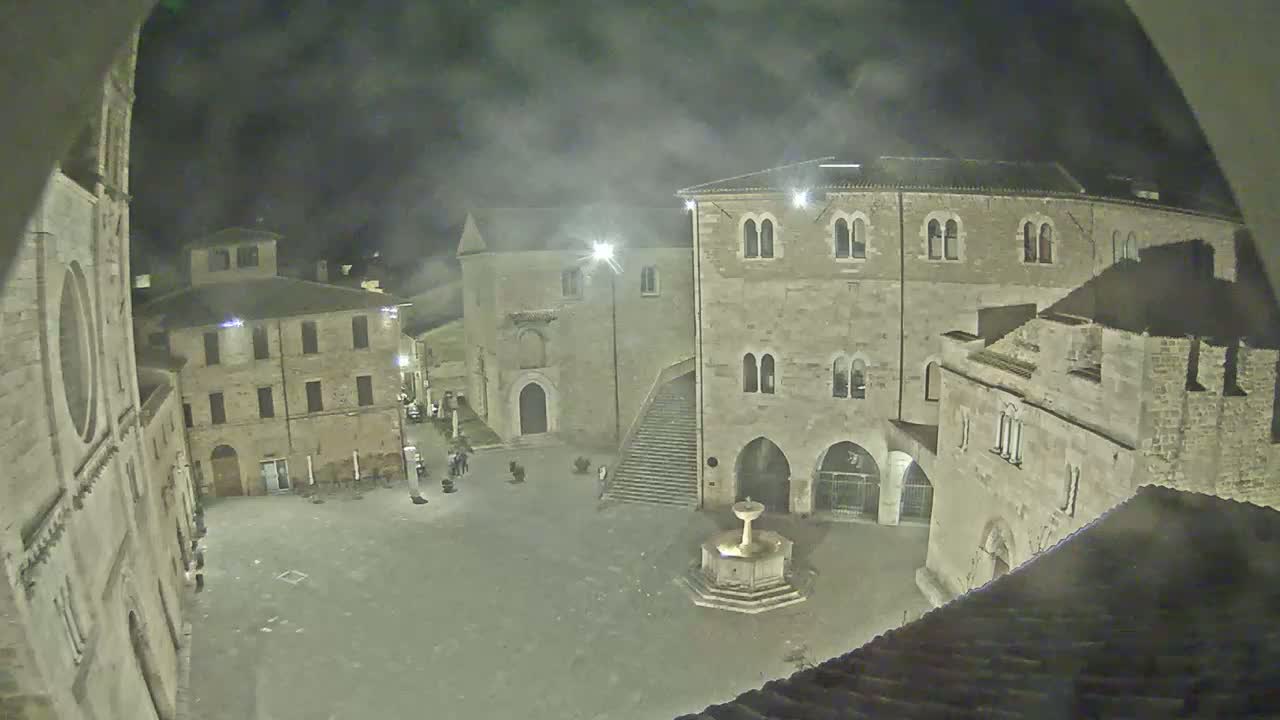 A nighttime view of a town square, surrounded by stone buildings and a fountain, under a cloudy sky.