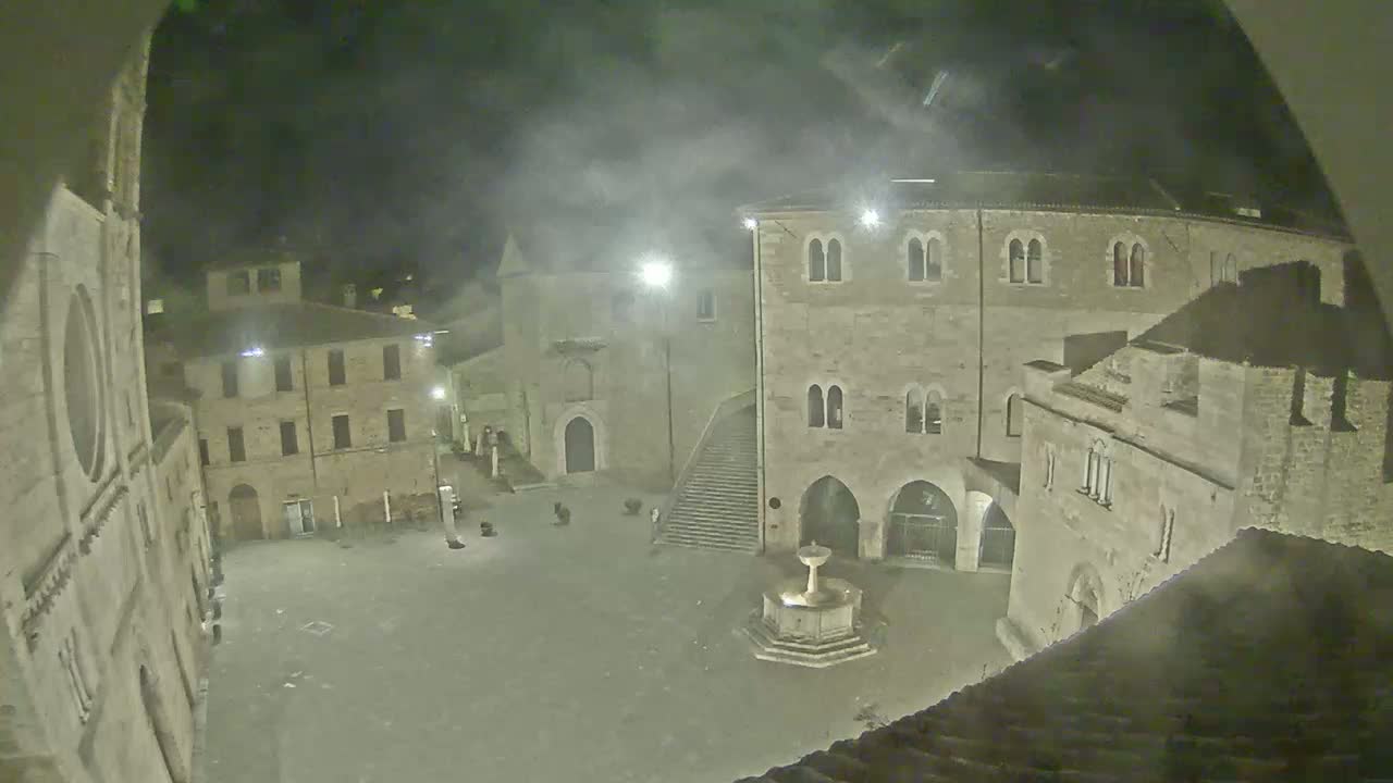 A nighttime view of a town square with stone buildings, a fountain, and hazy atmospheric conditions.