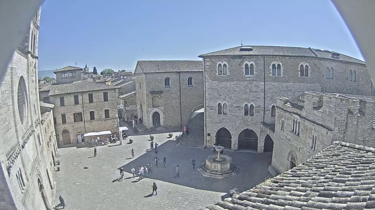A high-angle, sunny view of a town square with several stone buildings and a few people walking around, centered on a fountain.
