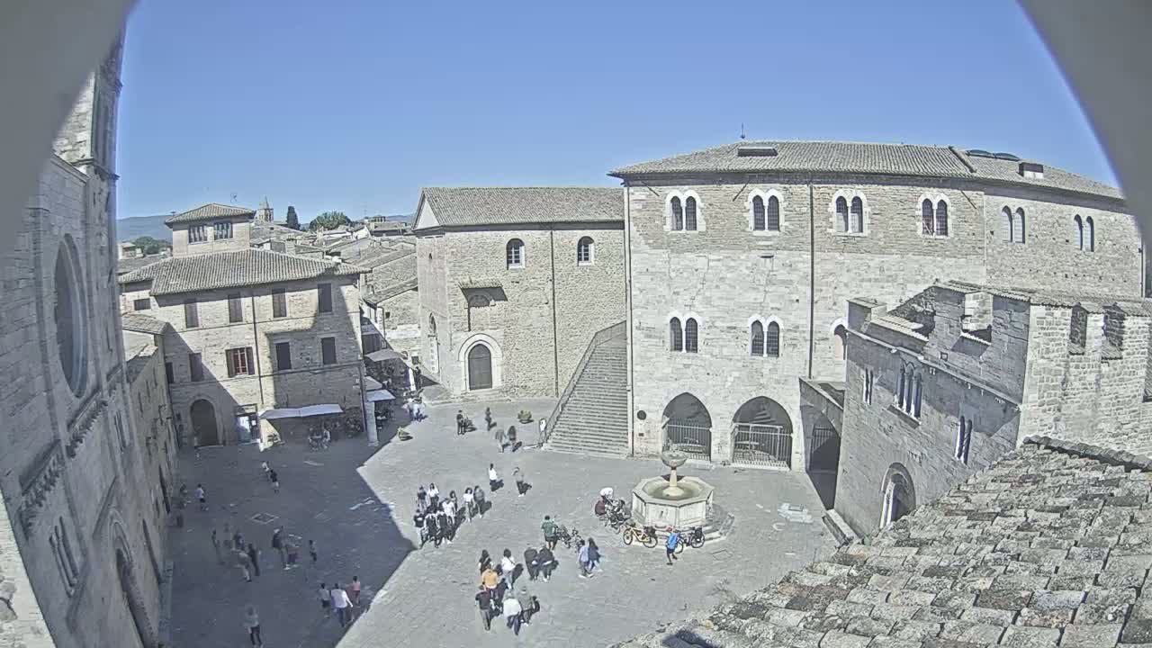 A sunny day reveals a town square surrounded by medieval stone buildings, with a fountain at its center and people walking about.
