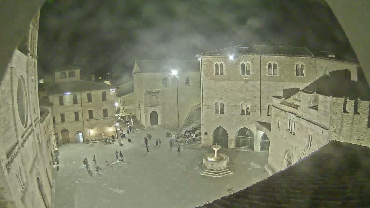 A nighttime, high-angle view shows a town square with several stone buildings surrounding a fountain, with a few people scattered throughout the square.
