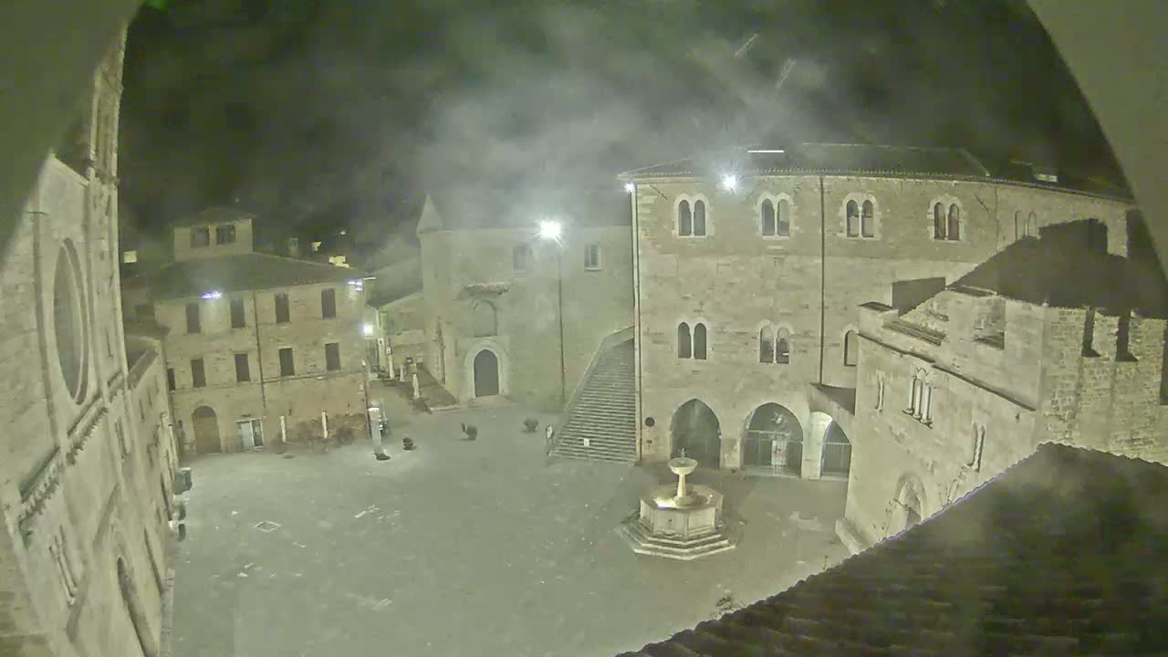 A nighttime view of a town square, surrounded by old stone buildings, with a fountain in the center and a hazy atmosphere.