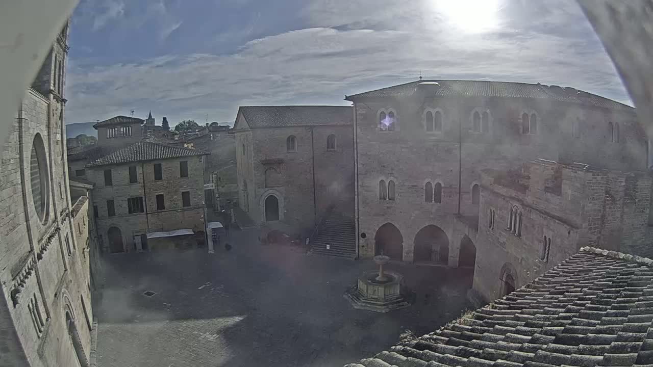 A sunny day reveals a town square surrounded by ancient stone buildings, one featuring a central fountain.