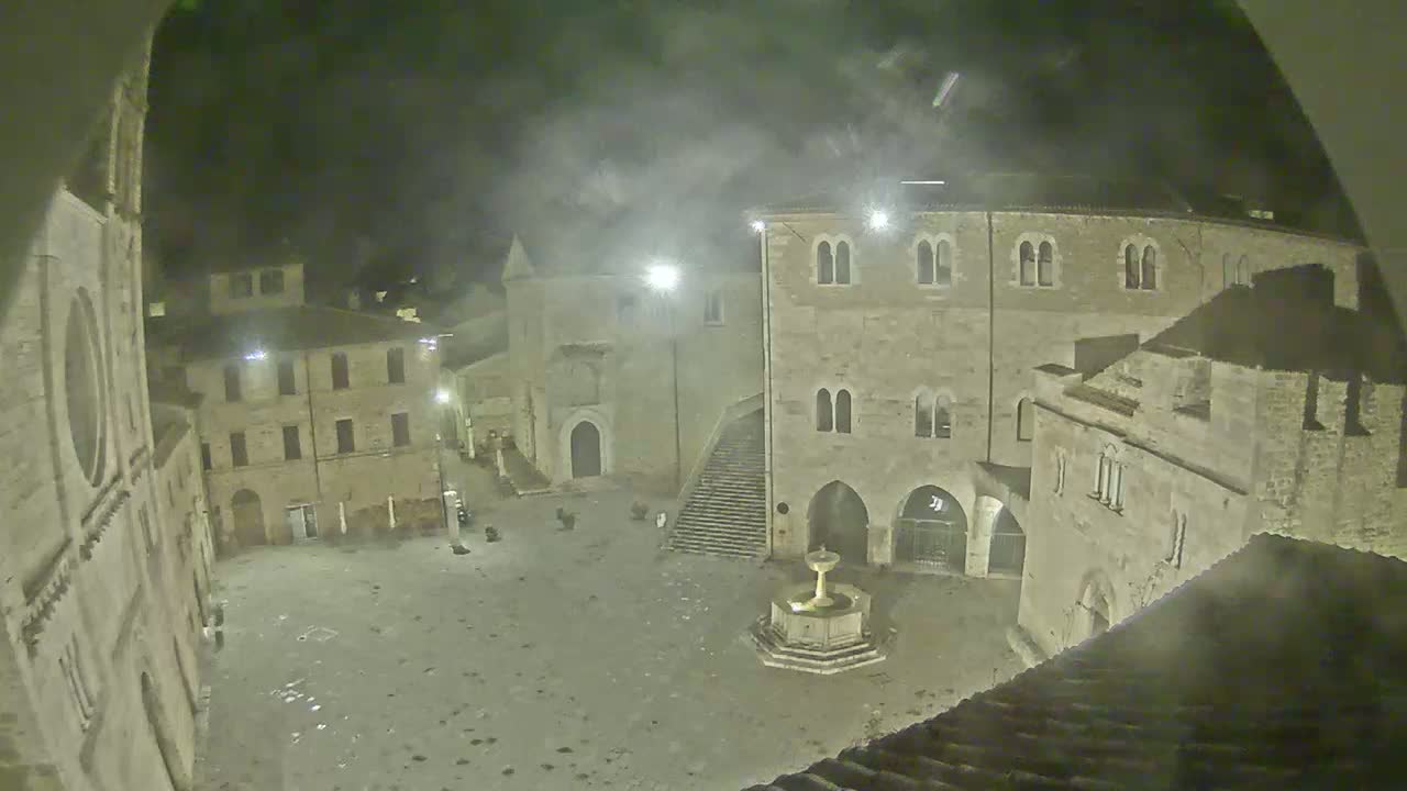 A nighttime view of a town square with stone buildings, a fountain, and hazy weather conditions.