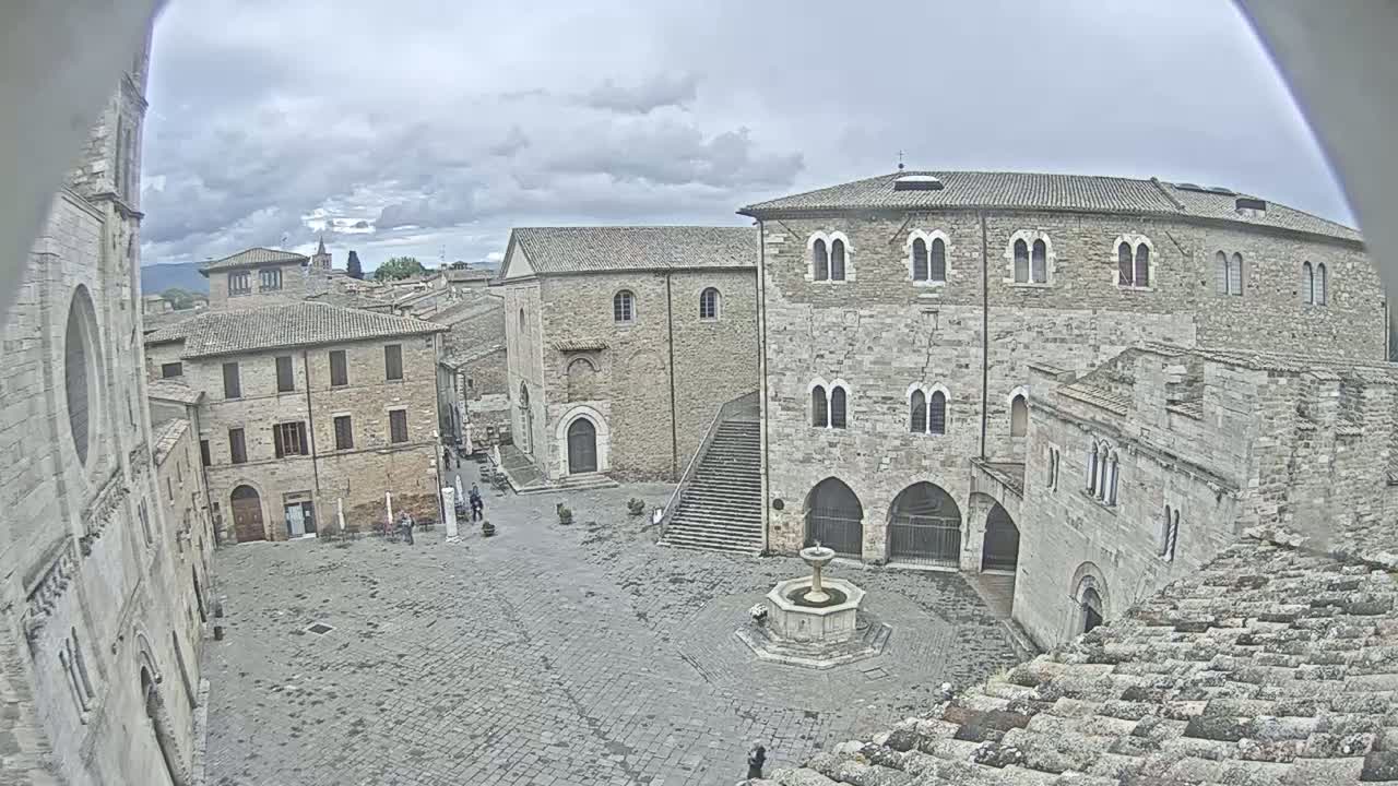 A stone plaza with a fountain is surrounded by medieval buildings under a cloudy sky.