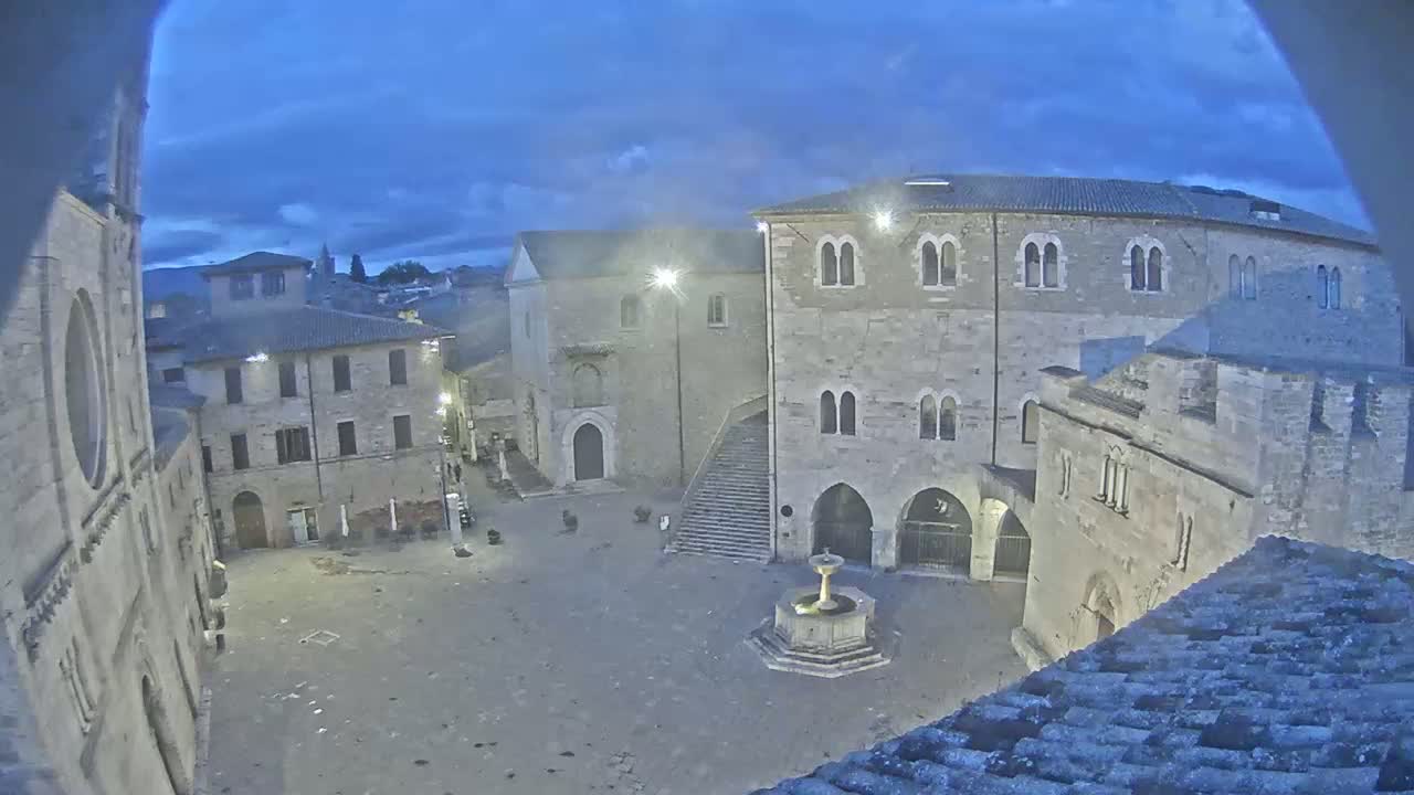 A stone plaza at twilight, centered around a fountain, is surrounded by ancient buildings under a mostly cloudy sky.