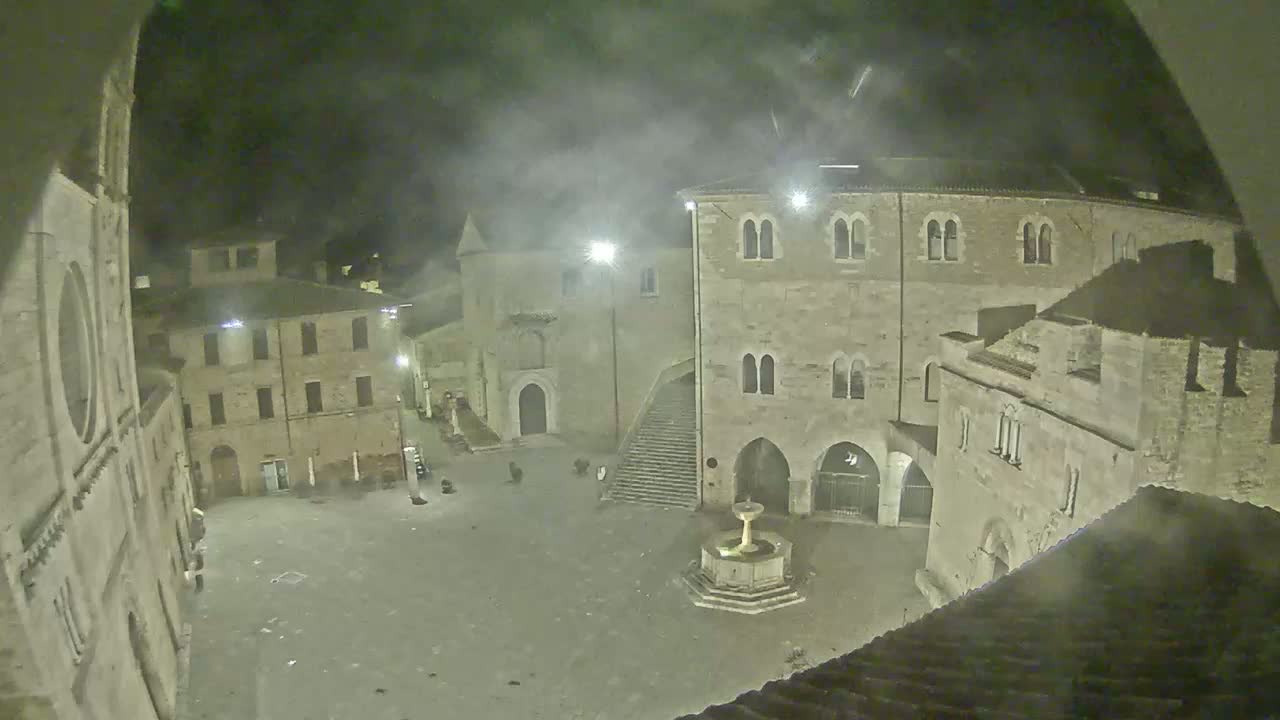 A nighttime view of a stone plaza in a small town, featuring several buildings and a fountain, under hazy or foggy conditions.