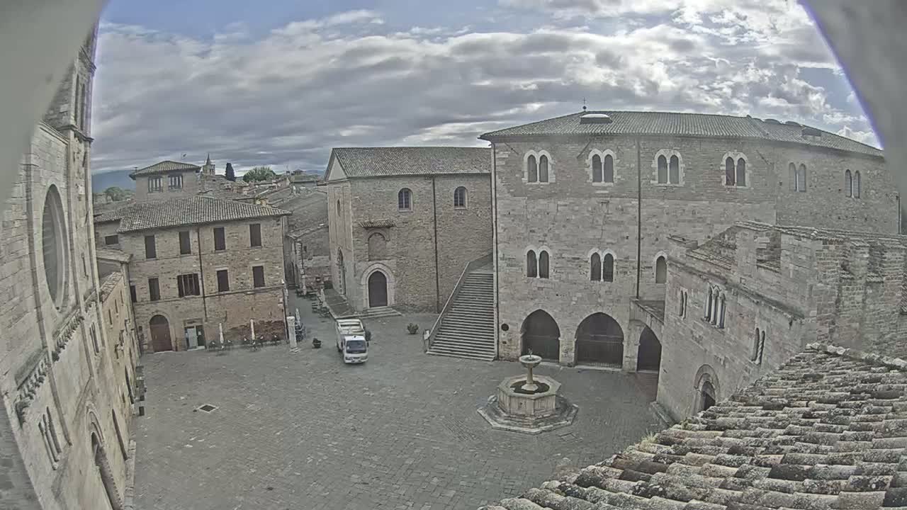 A high-angle view of a town square with stone buildings under a partly cloudy sky, featuring a fountain in the center and a single vehicle parked nearby.