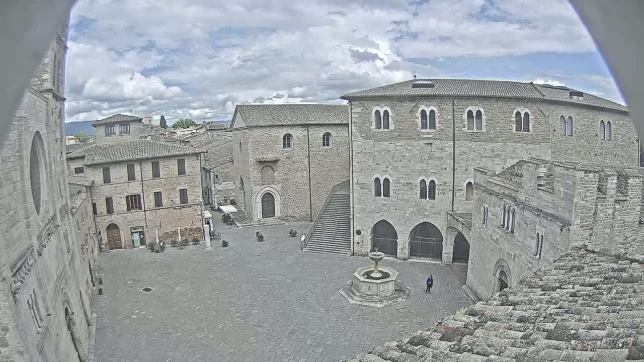 A stone plaza with a fountain in the center is surrounded by medieval-style buildings under a partly cloudy sky.