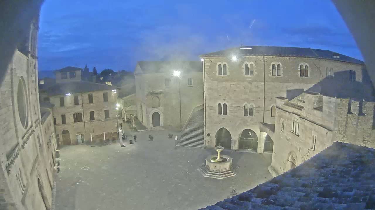 A nighttime view of a town square with stone buildings, a fountain, and a few people, under a twilight sky.