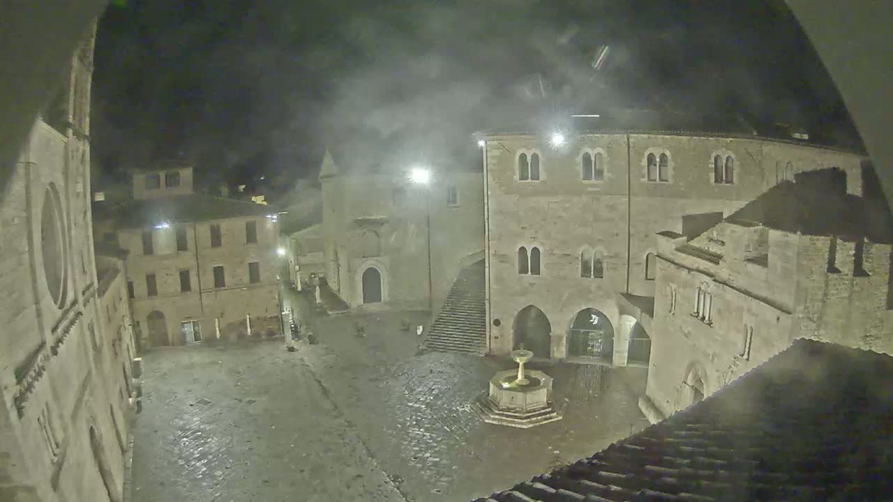 A nighttime, possibly rainy, view of a cobblestone town square surrounded by old stone buildings, a fountain at the center, and streetlights illuminating the scene.