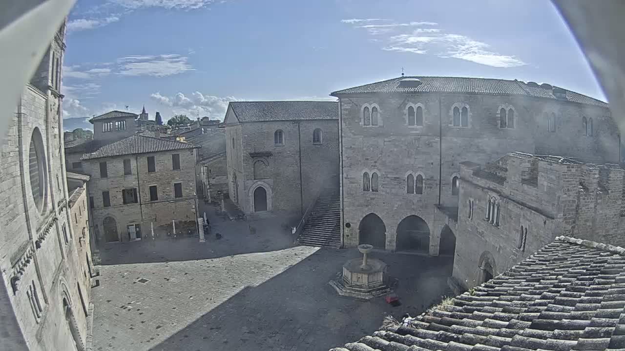 A partly sunny day reveals a large stone plaza surrounded by historic buildings, including a fountain in the center.