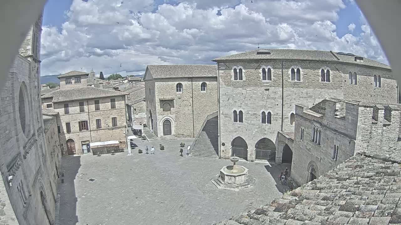 A partly cloudy day reveals a town square in a European-style village, centered on a stone fountain, surrounded by aged stone buildings.