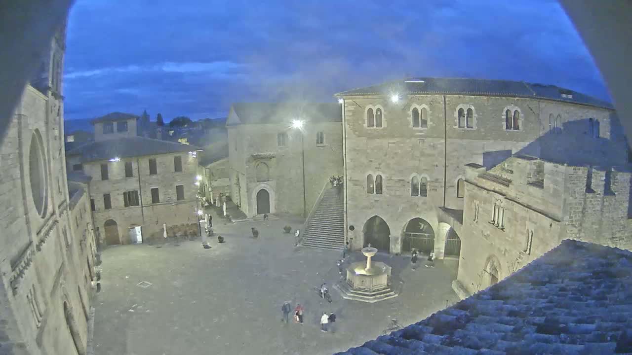 A twilight view of a stone town square with a fountain, surrounded by medieval-style buildings.