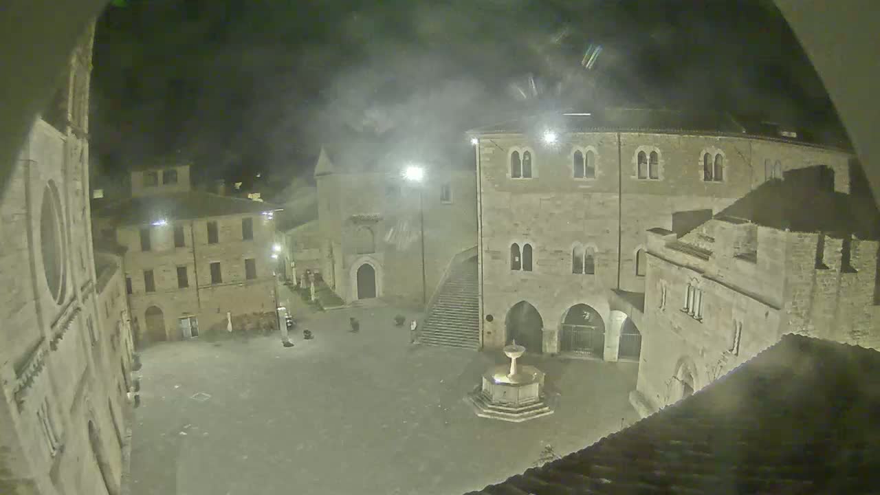 A nighttime view of a town square, featuring stone buildings and a fountain, under hazy or foggy conditions.