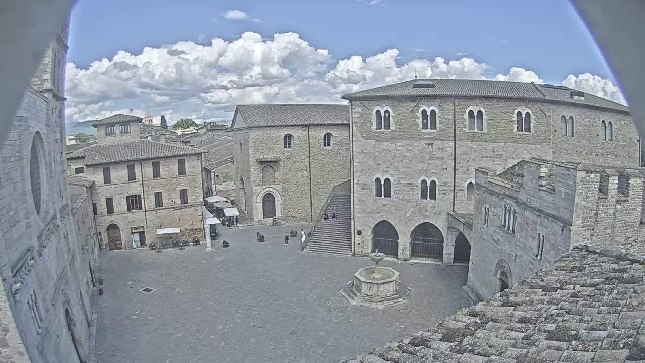 A partly cloudy day reveals a large stone plaza surrounded by medieval buildings, including a central fountain and a set of stairs.
