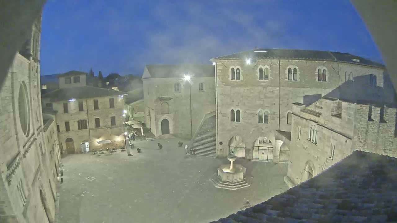 A stone town square at night is sparsely populated with a fountain at its center, surrounded by old buildings under a twilight sky.