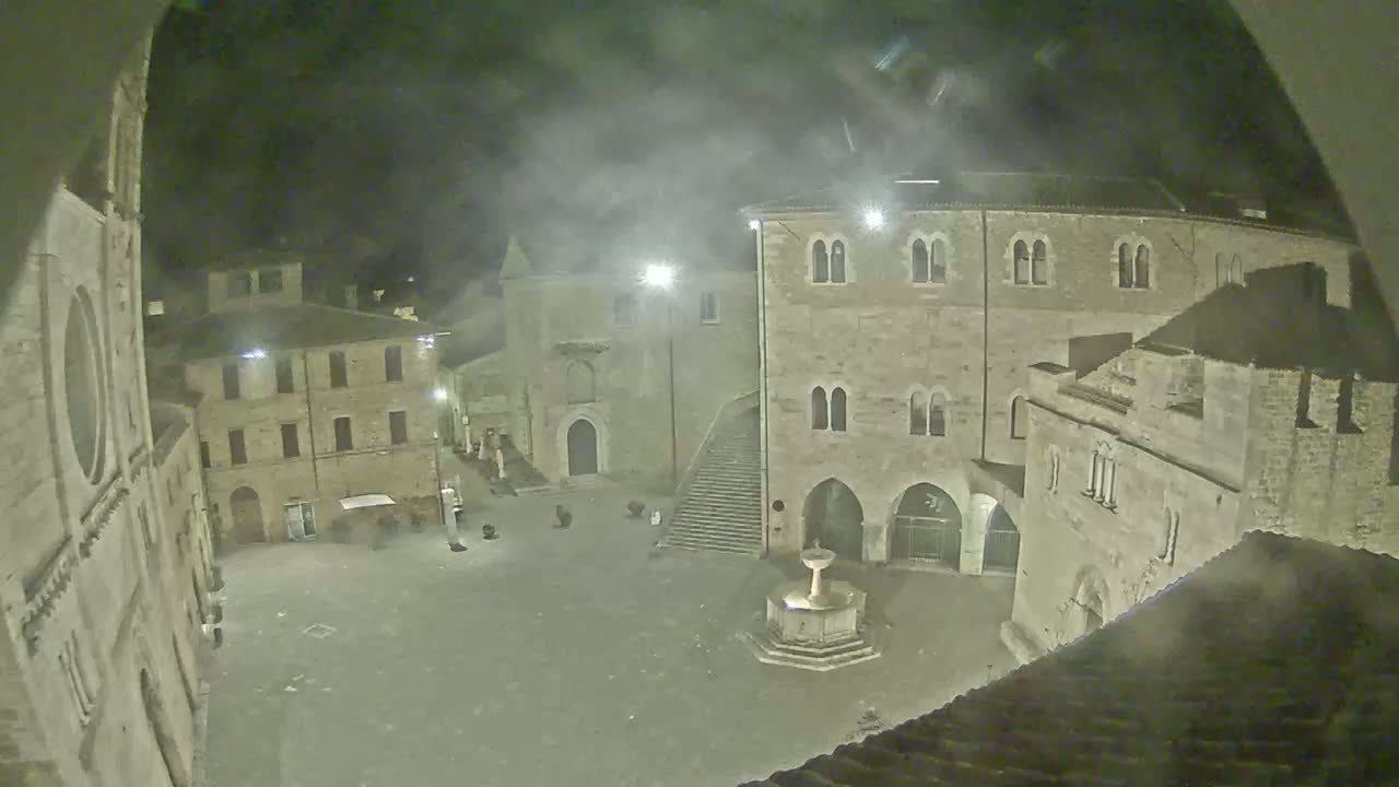 A nighttime view of a town square, with old stone buildings surrounding a fountain, under hazy or foggy conditions.
