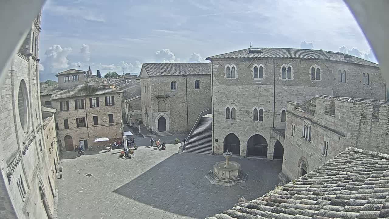 A sunny day overlooks a town square with several stone buildings, a fountain, and a few people.