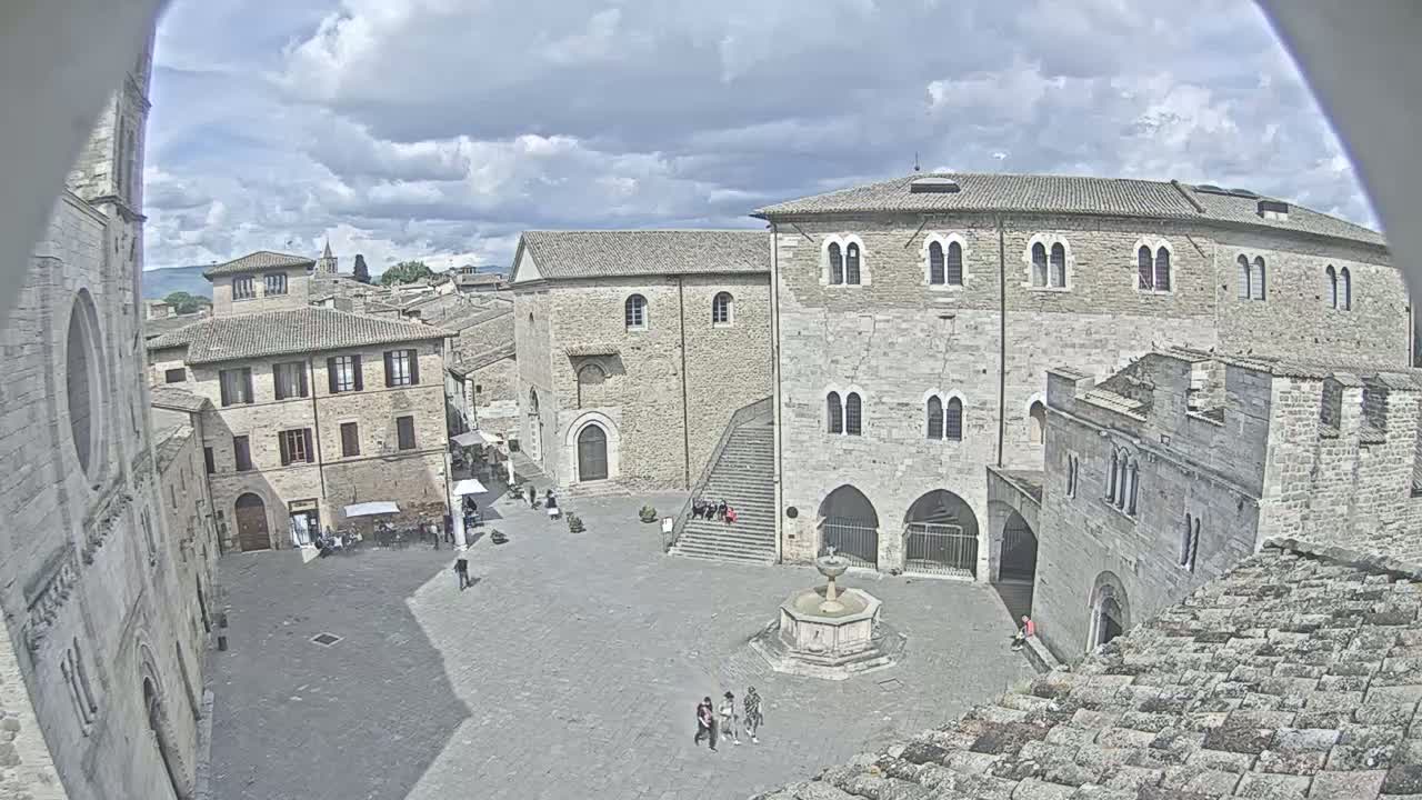A partly cloudy day reveals a high-angle view of a town square with several stone buildings, a fountain, and a few people walking around.