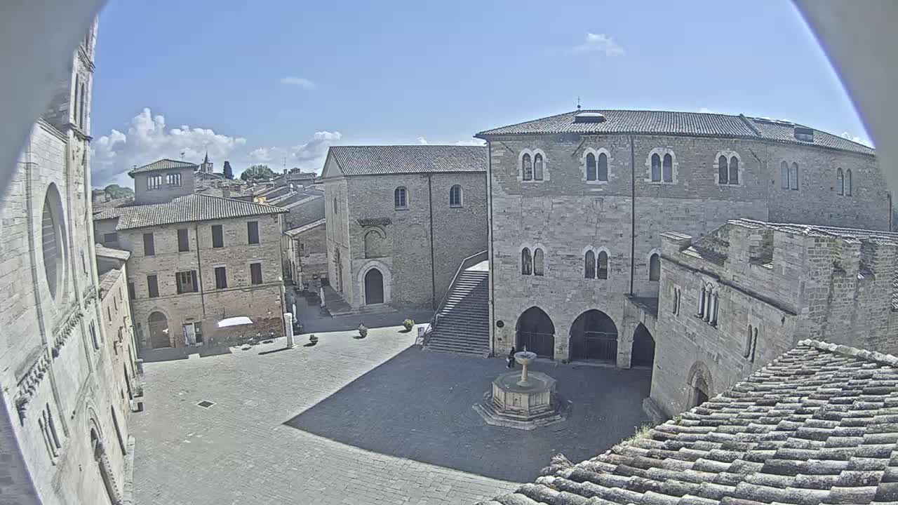 A sunny view of a town square in Italy, featuring ancient stone buildings surrounding a fountain, with a partly cloudy sky above.