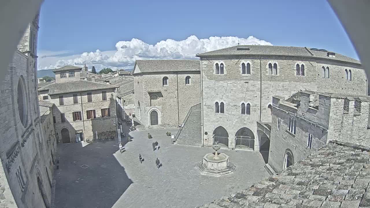 A mostly sunny day reveals a large, stone plaza in a historic European town, centered around a fountain, with several people walking and buildings surrounding the square.