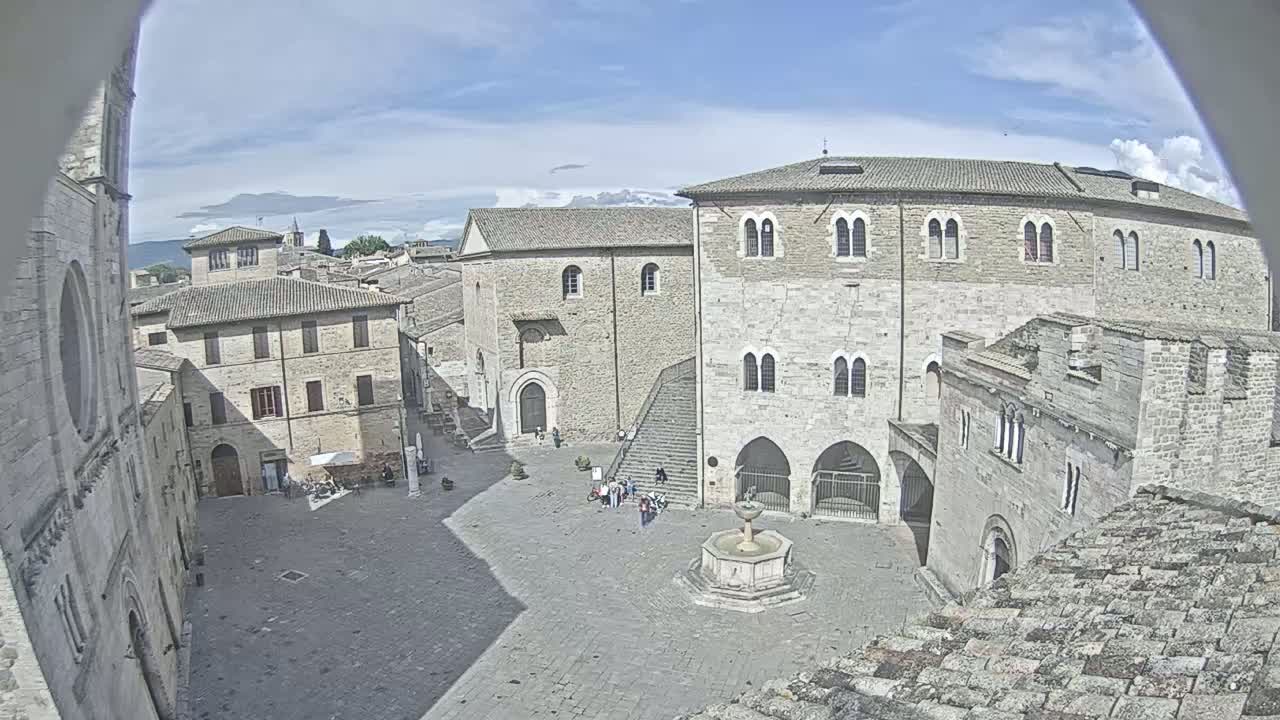 A sunny day reveals a high-angle view of a town square in Italy, surrounded by ancient stone buildings, including a large, multi-storied structure, with a few people strolling near a central fountain.
