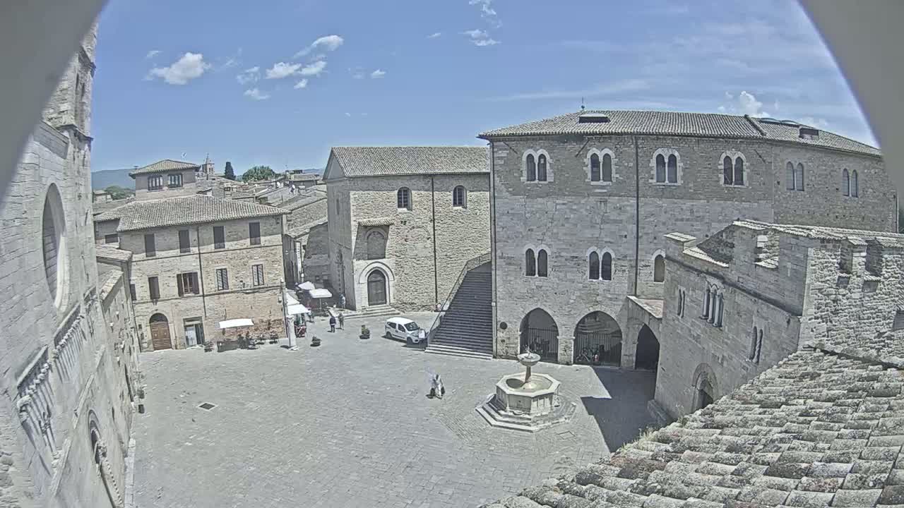 A stone plaza, under a mostly sunny sky, is surrounded by aged buildings, one with a fountain in the center.