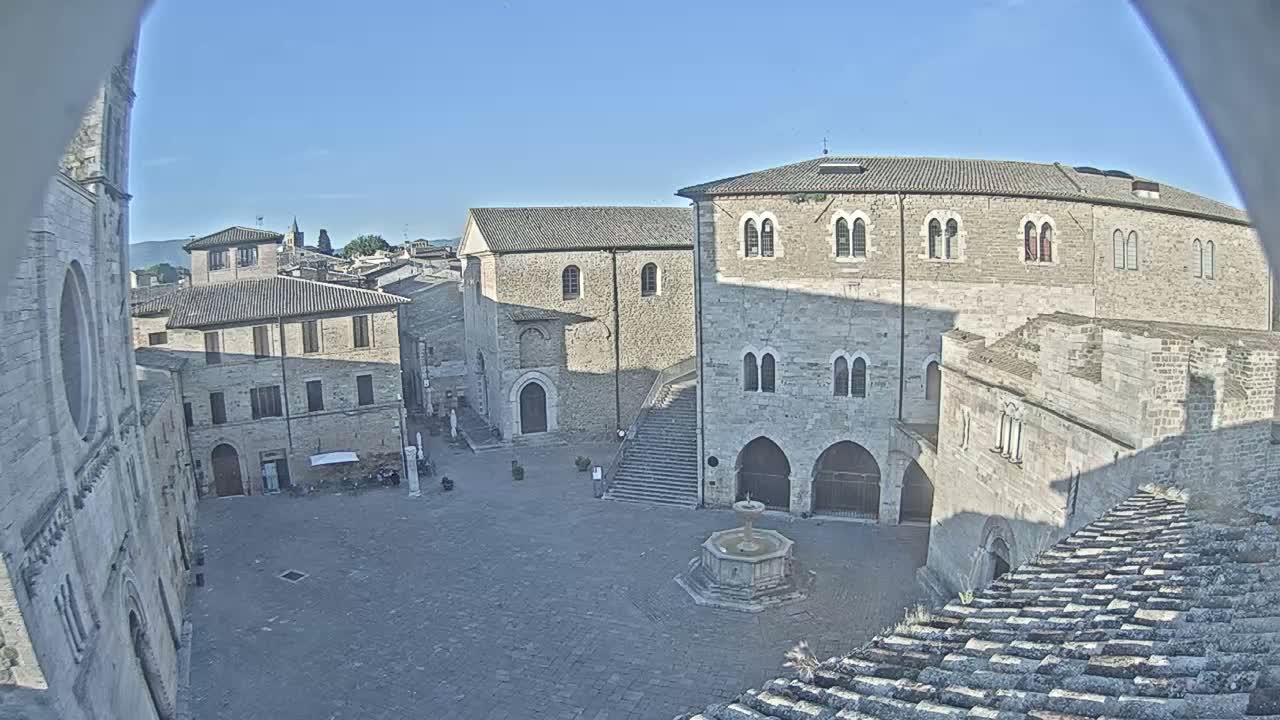 A sunlit town square, featuring several stone buildings and a central fountain, is viewed from an elevated perspective on a clear day.