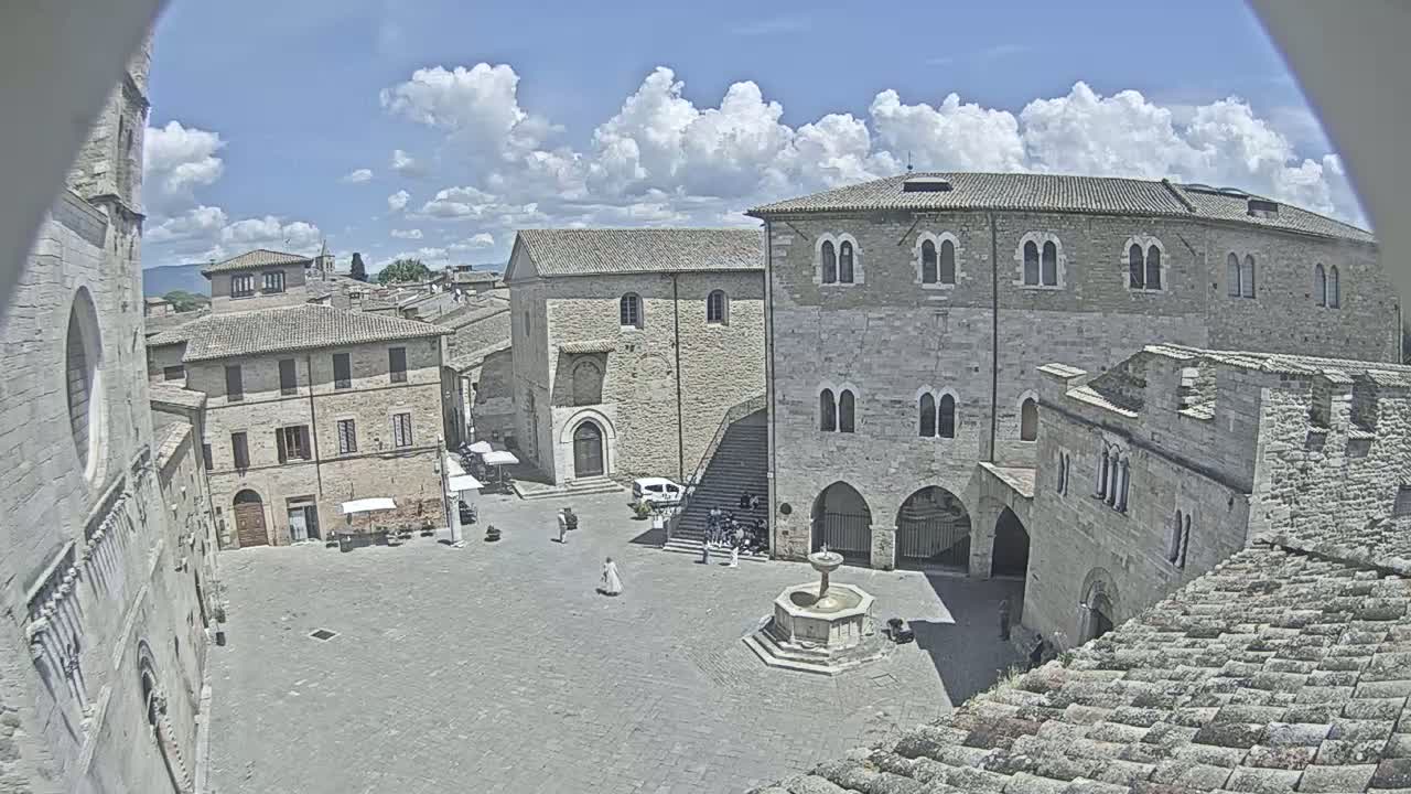 A stone plaza with a fountain is surrounded by medieval-looking buildings under a partly cloudy sky.