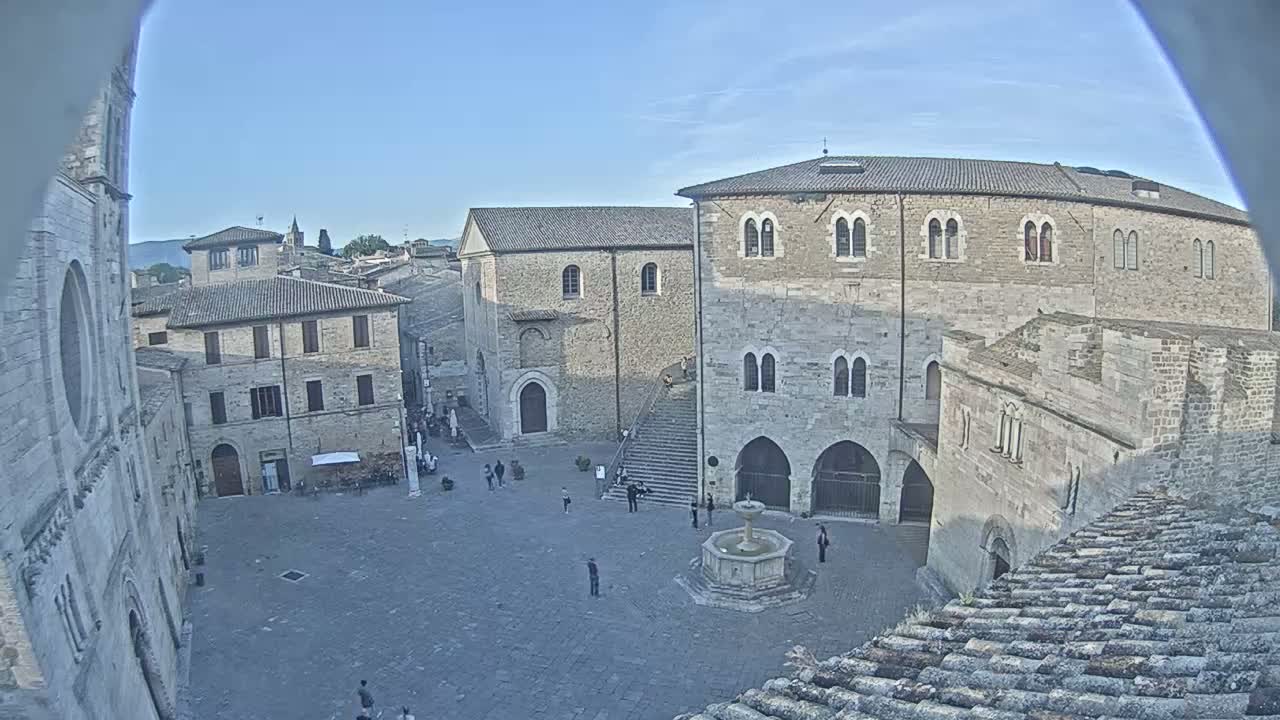 A stone plaza with a fountain in the center is surrounded by several historic stone buildings under a clear blue sky.