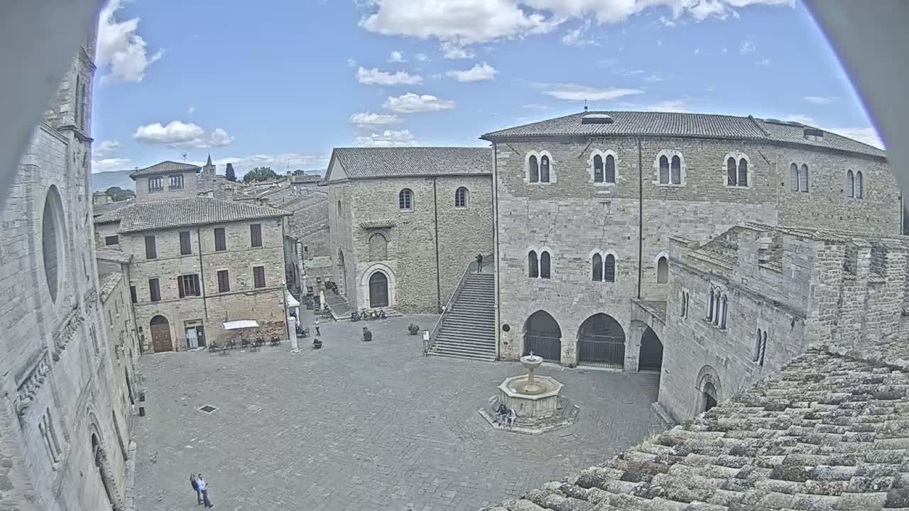 A mostly sunny day reveals a large town square surrounded by stone buildings, a central fountain, and a stairway leading up to one of the structures.