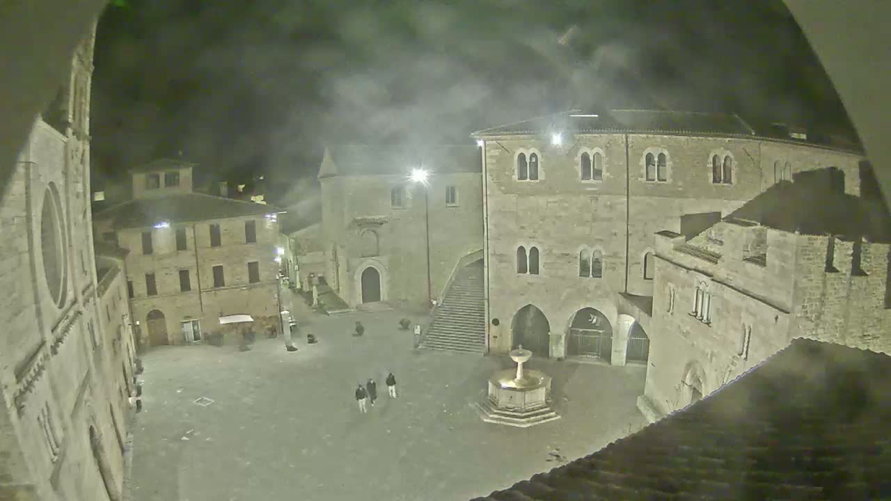 A nighttime view of a town square with several stone buildings surrounding a fountain, under hazy or foggy conditions.