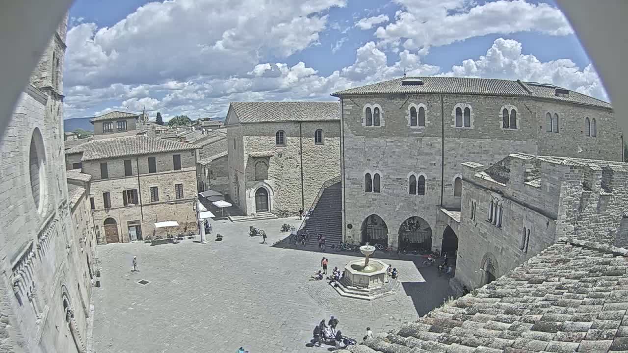 A partly cloudy day reveals a town square surrounded by medieval stone buildings, one with a prominent fountain at its center.