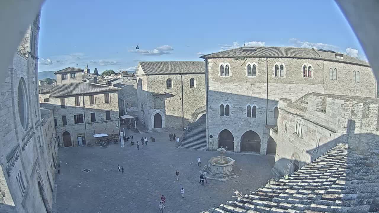 A mostly empty, stone plaza with a fountain is surrounded by old, light-colored buildings under a clear blue sky.