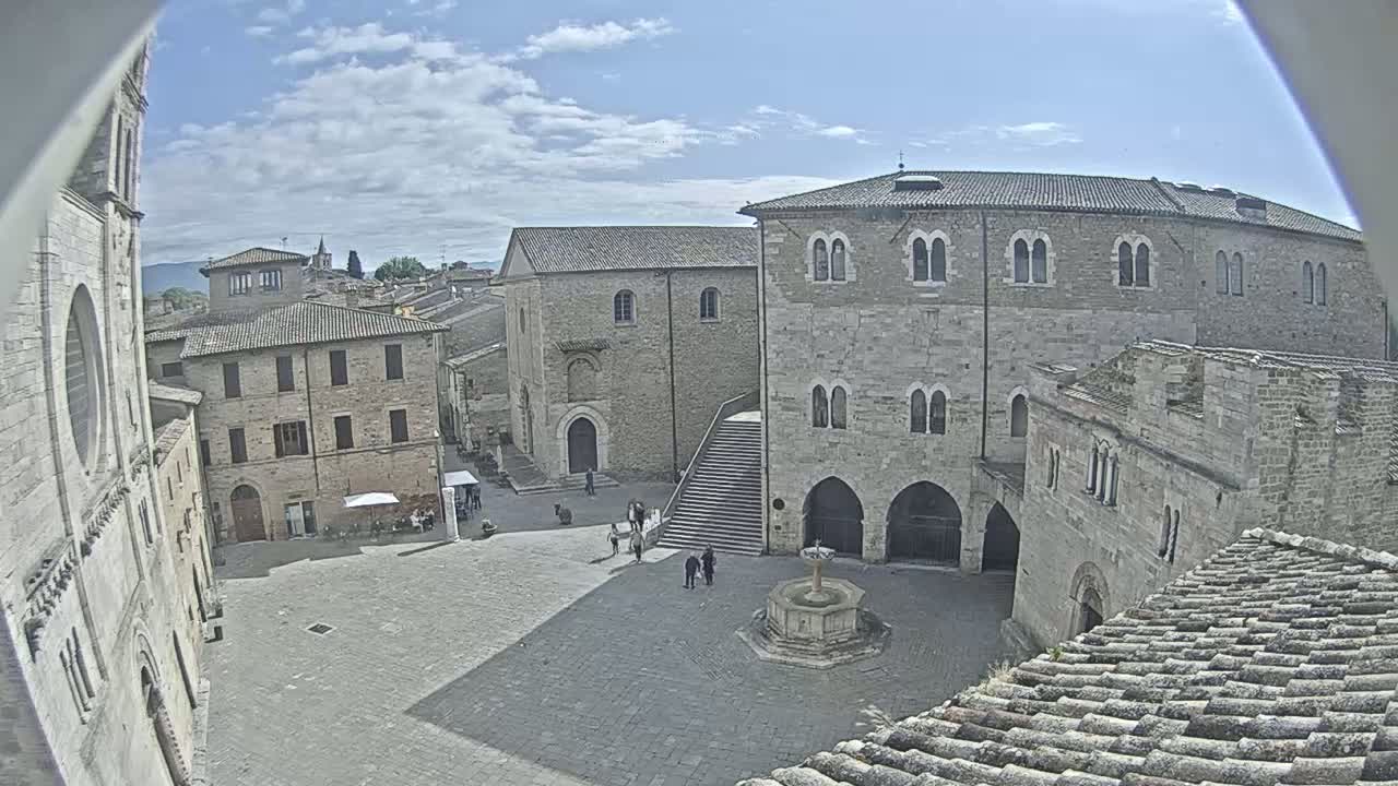 A mostly sunny view of a town square surrounded by stone buildings, including a large church and a multi-story structure with a fountain in the center.