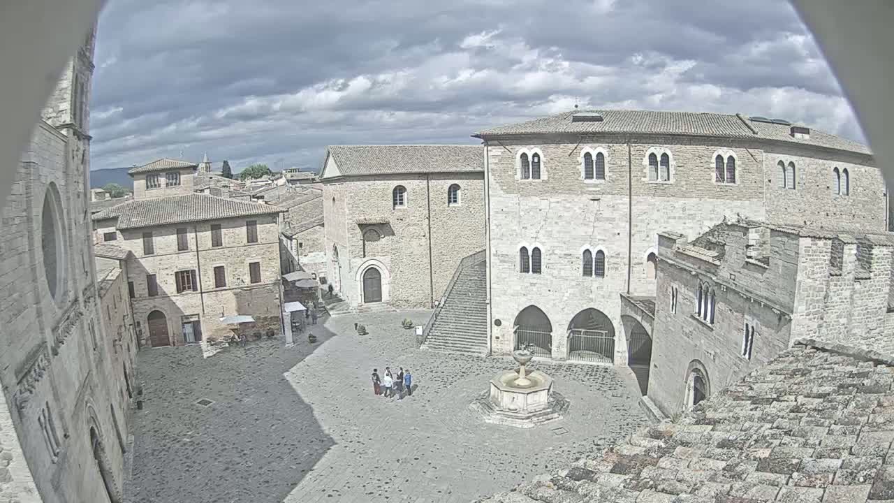 A high-angle view of a town square with several stone buildings under a cloudy sky, showing a fountain and a small group of people.