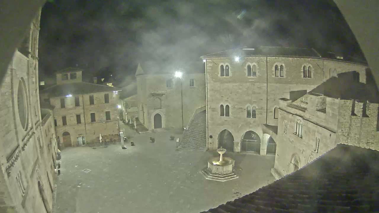 A nighttime view of a stone plaza in a historic town, possibly under a hazy or foggy sky, features buildings with arched entryways and a central fountain.