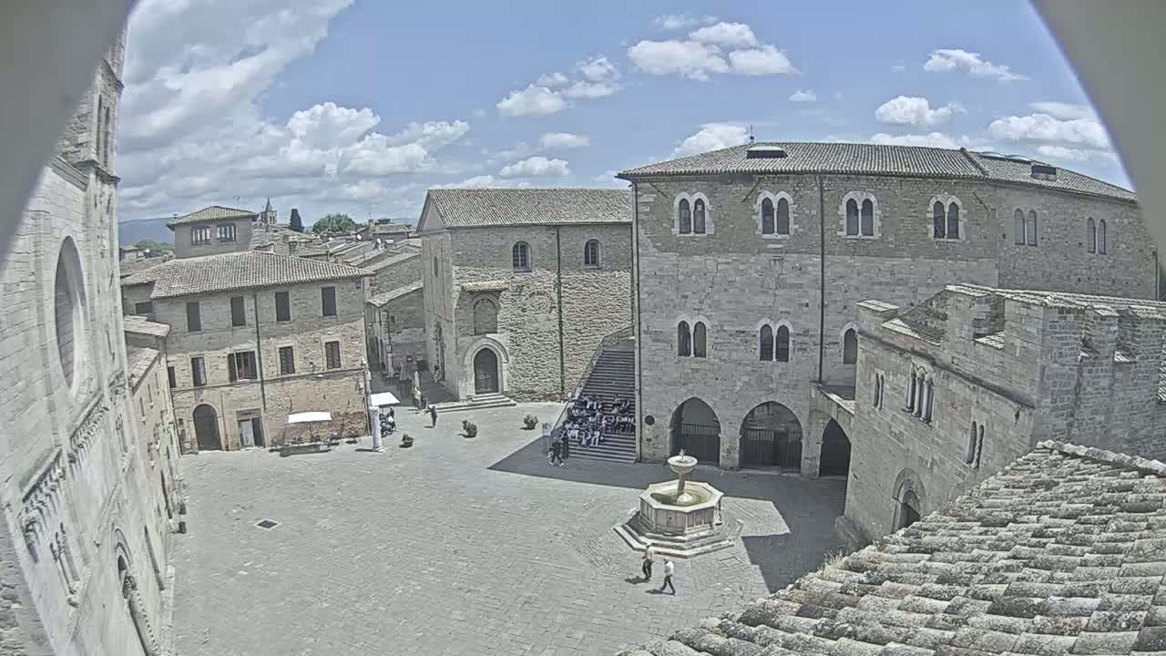 A mostly sunny day overlooks a town square with several stone buildings, a fountain, and a few people.